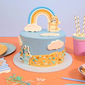 Bluey Cake by Crumbs & Doilies with Bluey and Bingo edible toppers and rainbow on blue icing.